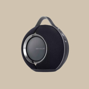 Devialet Mania, en elegant svart bärbar högtalare med metalldetaljer och ett praktiskt handtag, står redo mot en beige bakgrund.