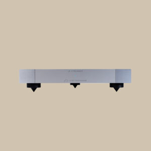 En elegant silverfärgad Le Streamer står redo att förhöja ljudet, med Metronome-logotypen lysande, balanserad på tre koniska fötter mot en beige scen.