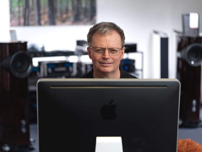 En man med glasögon sitter vid sin stora Apple-skärm, omgiven av premiumhögtalare och ljudutrustning, skogskonst suddig bakom honom.