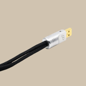 Pachanko Nano Silver USB 1,5 m-kabeln med sin svarta flätade hölje, silverfärgade kontakt och guldfärgade sticka, som verkligen sticker ut mot den beige bakgrunden.