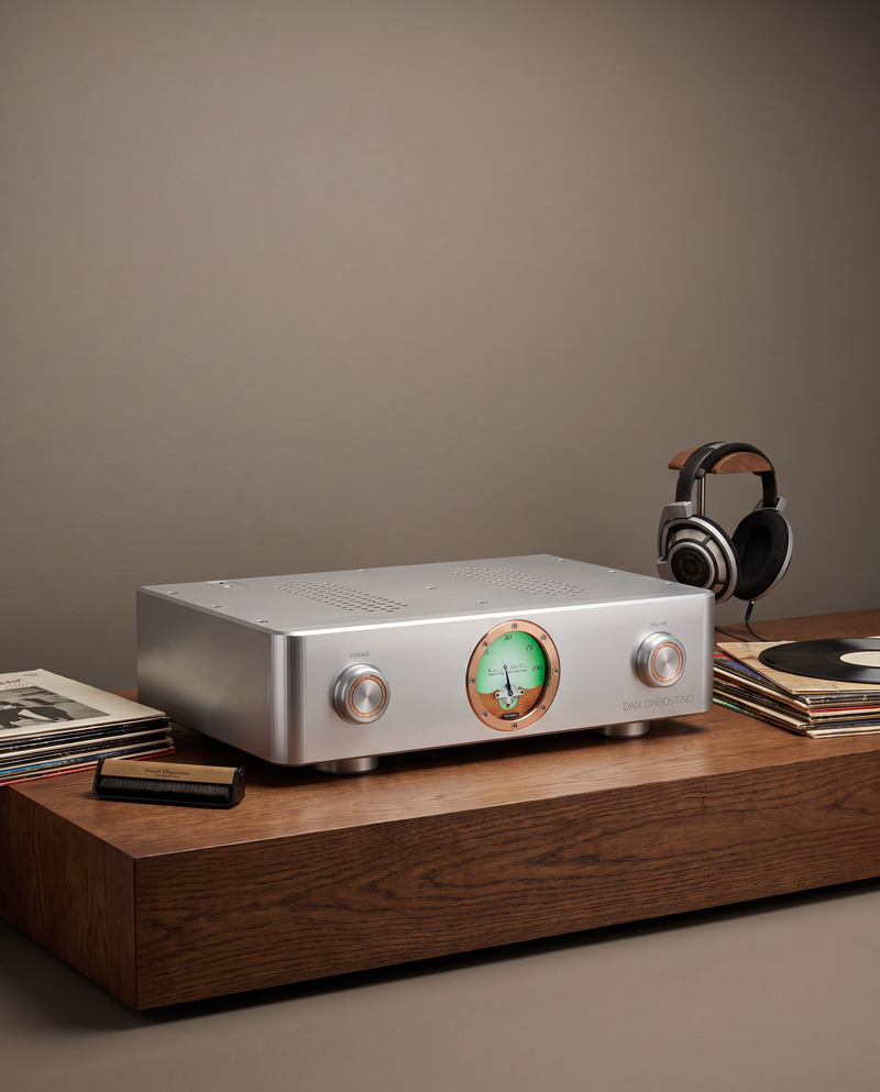 En elegant silverfärgad ljudförstärkare står på ett varmt träbord, omgiven av hörlurar som är redo för djupgående lyssningsstunder, klassiska vinylskivor som väntar på att spelas och en stämningsfull munspel. På frontpanelen pryder en stor analog mätare och två knappar med tydlig taktil känsla – som inbjuder till praktisk reglering för att uppnå det perfekta ljudet.